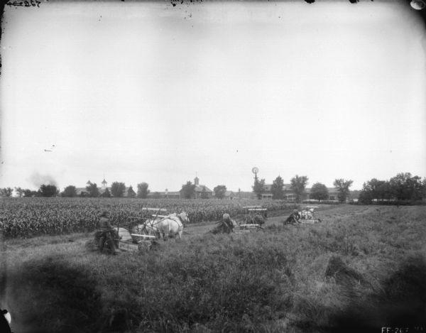 Slightly elevated view of three men using horse-drawn binders. Farm buildings are in the background.