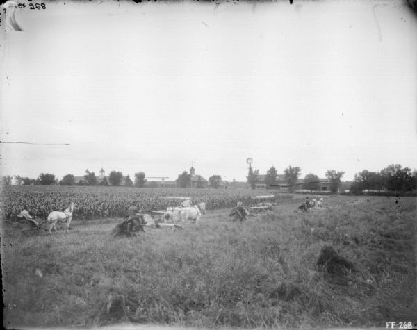 Slightly elevated view of three men using horse-drawn binders. Another man rides in a horse-drawn buggy behind the binder on the left. Farm buildings and a windmill are in the background.
