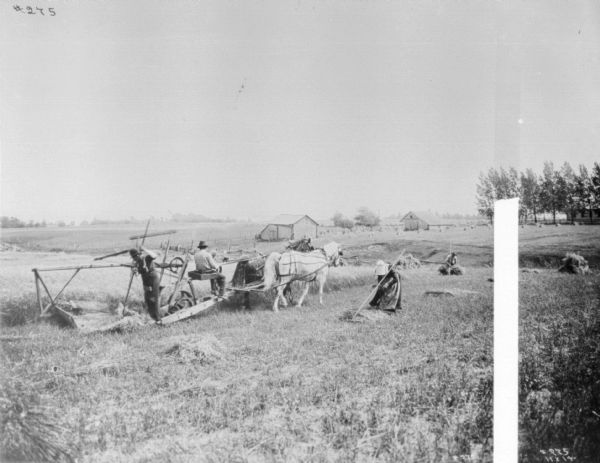 View across field towards a man using a horse-drawn reaping and mowing machine. Another man is standing behind the machine raking up the grain. A woman is working in the field in the center near the binder, and another man is working in the field behind on a slope. Farm buildings are in the background.