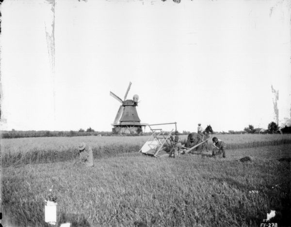 View across field towards people working with harvested grain near a reaping and rowing machine. A windmill is in the background. Farm buildings are in the background on the right.
