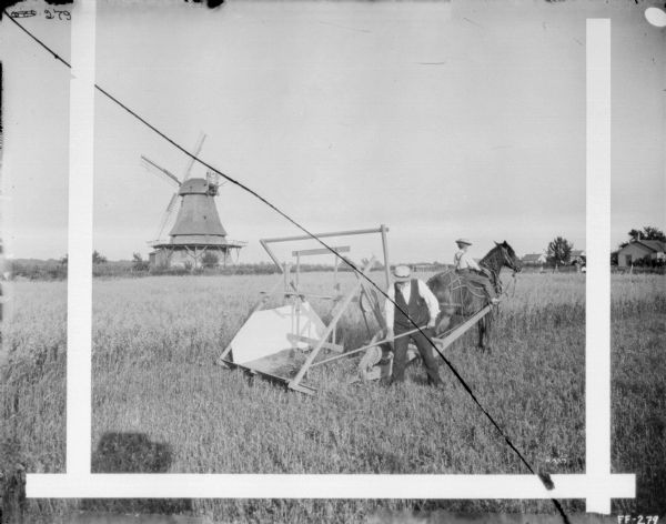 A man and a young boy are using a reaping and mowing machine in  field. There is a windmill in the background on the left, and farm buildings are on the right.