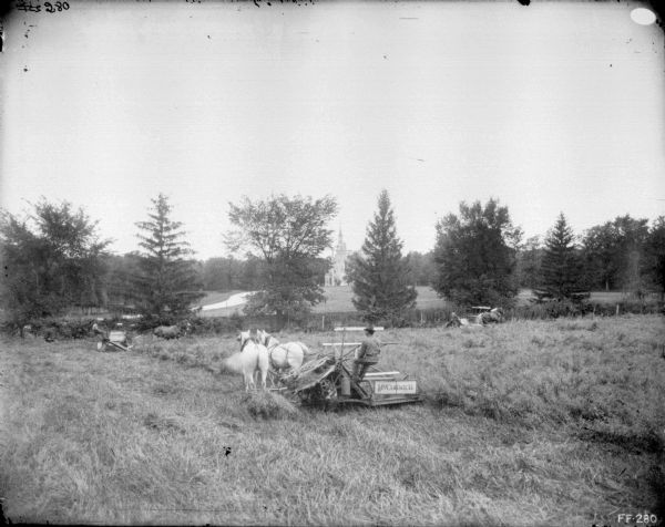 Three men using McCormick horse-drawn binders in a field. A road leading to a large building with a turret is in the background.