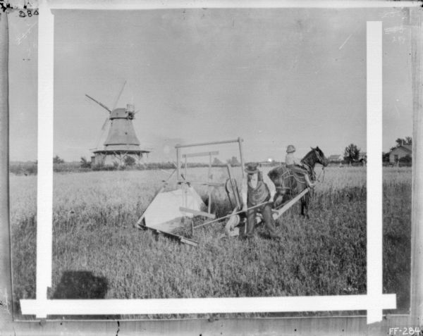 Slightly elevated view of a man and a young boy who are using a reaping and mowing machine. The boy is sitting on the horse pulling the machine. There is a windmill in the background on the left, and farm buildings on the right.
