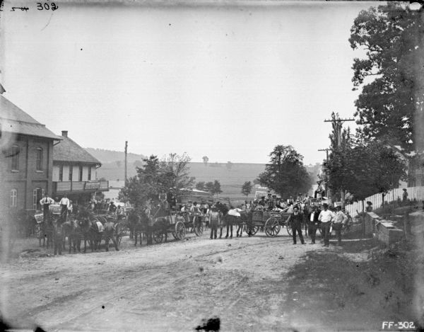 View down unpaved road towards a large group of people, mostly men, with some children, gathered on and around horse-drawn wagons near commercial buildings. McCormick signs are on the side of some of the wagons, and some of the men hold McCormick signs, as well as posters. In the background the road rises up a steep hill between fields and farm buildings.