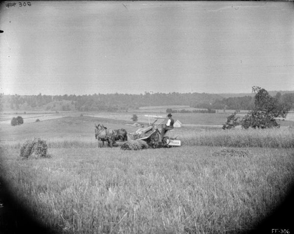 View down low hill towards a man using a horse-drawn McCormick binder in a field. In the far background are more fields, and a tree-lined hill.