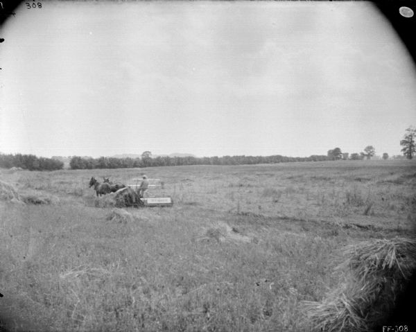 View across field towards a man using a horse-drawn McCormick binder in a field.