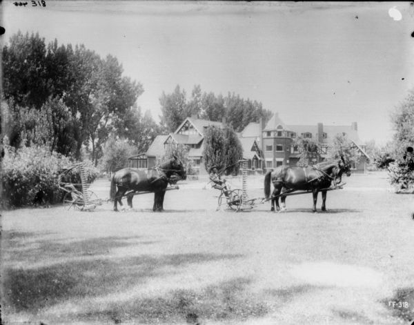 View across lawn towards two men posing with horse-drawn mowers on the lawn of an estate. A mansion is in the background among trees and shrubs.