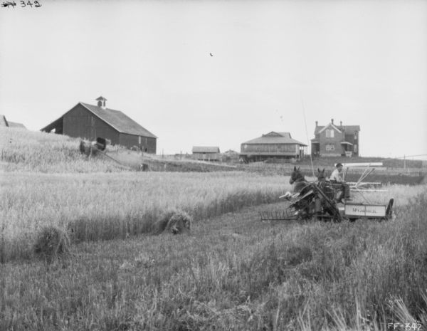 View of a man using a horse-drawn McCormick mower in a field. A barn and other farm buildings are in the background on a low hill.