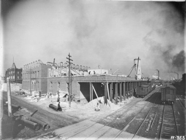 Slightly elevated view of a twine mill under construction. Railroad cars are on railroad tracks running along the buildings under construction. Men are working on the building in the foreground. Other workers are on the roof.