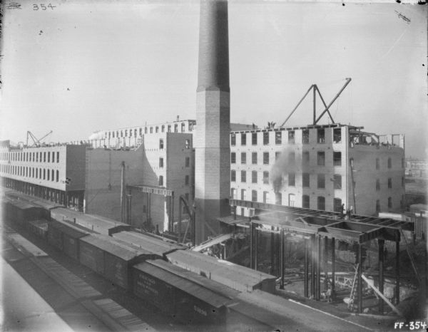 Elevated view of a twine mill under construction. Railroad cars are on tracks in the foreground. A large smokestack is in the center.