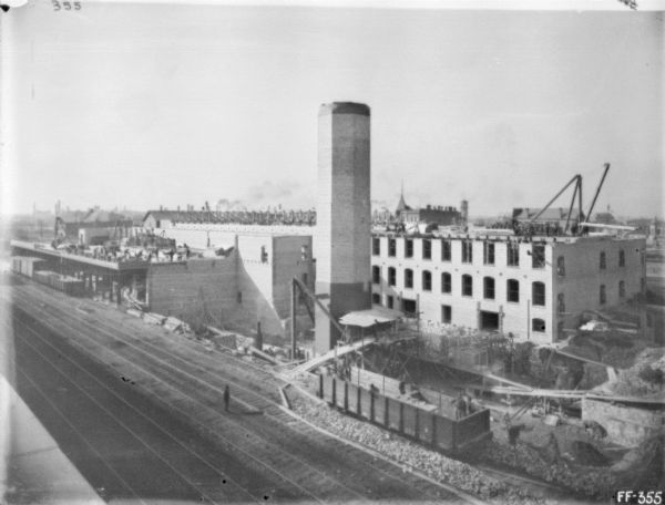 Elevated view of a twine mill under construction. Railroad tracks are in the foreground below. A partially built smokestack is in the center. A number of construction workers are on two different roof sections.