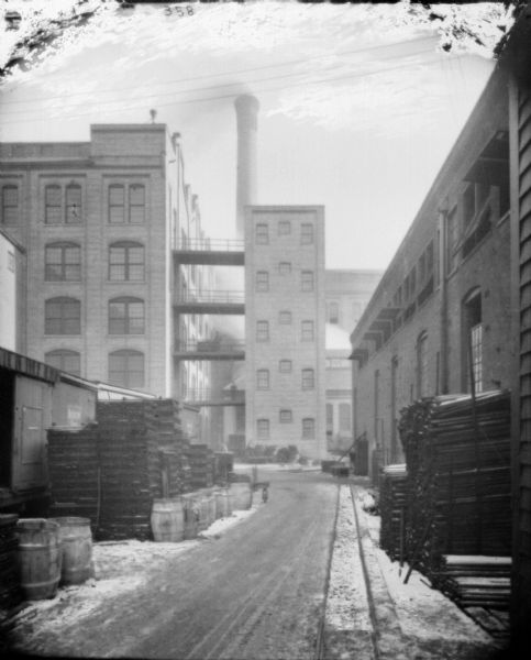 View down alley towards lumber storage in plant yard near railroad cars. Brick factory buildings are in the background. Walkways on the upper floors link two buildings. There is a large smokestack in the background.