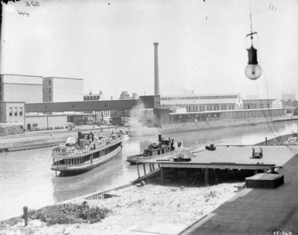 Slightly elevated view from shoreline towards the McCormick Works on the opposite shoreline. There is a boat in the river near a tugboat. There is a light bulb hanging from a wire in the right foreground.