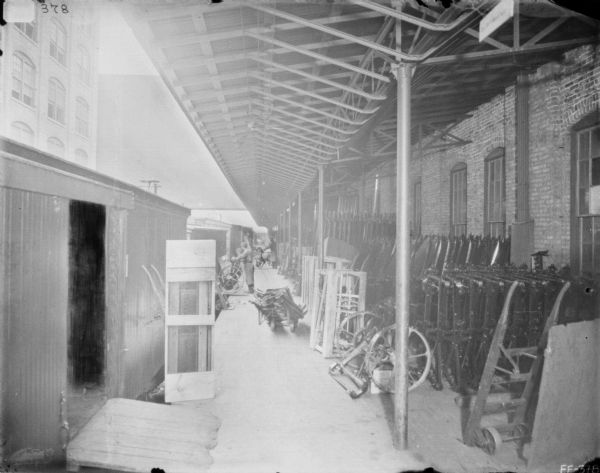 View down covered loading dock at McCormick Works. Men are loading or unloading machinery parts and are standing near railroad cars. Parts are lined up along the brick factory wall under the roof on the right. A brick factory building is on the left.