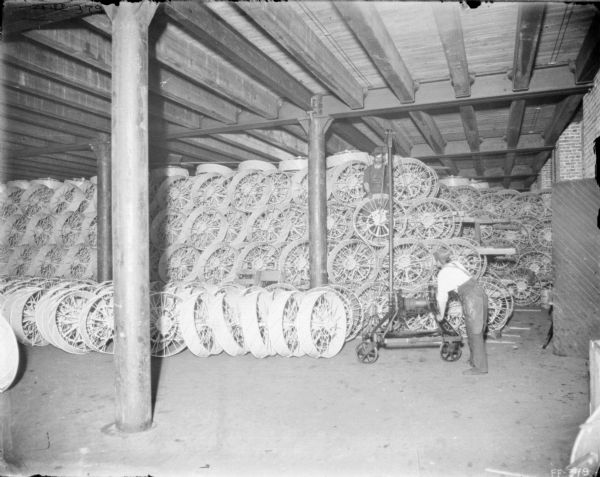 Interior view of men in the main wheel room of McCormick Works. One man is standing with a piece of machinery near the stacks of wheels which is probably for transporting the heavy wheels. There is another man holding one of the wheels while standing up on the stacks of wheels.