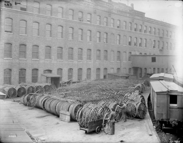 Elevated view of plant yard. Stacks of wheels are in rows in the center of the yard on a wood platform, and along the brick wall of the factory building in the background. Two men, one with a cart, stand near the wheels, and railroad cars on railroad tracks are on the right. On the facade near the roof of the factory building is a sign that reads: "Reaper Manufactory."