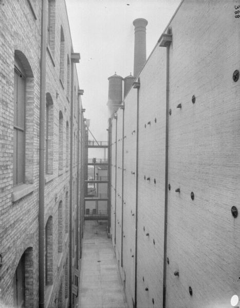 Elevated view of alley between two brick factory buildings at McCormick Works. In the background are four open walkways linking the two buildings. Two water towers and a chimney are behind the building on the right.