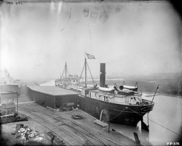 Elevated view of ship at a dock. Railroad tracks are in the foreground near low factory and storage buildings. The name on the back of the ship reads: "Northwestern Fairport." In the far background is a bridge.