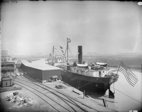 Elevated view of ship at a dock. Railroad tracks are in the foreground near low factory and storage buildings. The name on the back of the ship reads: "Northman Fairport." In the far background is a bridge.