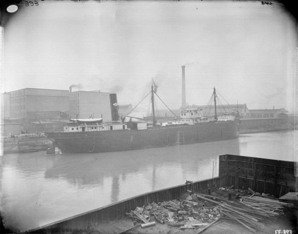 View across water towards ship, the "Northman Fairport," at a dock at McCormick Works. Factory buildings are in the background. In the foreground is a walled pier or storage area with metal or wood parts in piles.