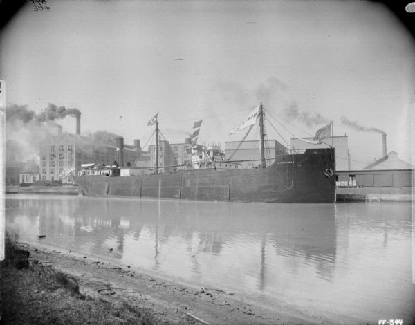 View from shoreline across water towards opposite shoreline where a ship is at the dock of McCormick Works. Factory buildings are in the background near smokestacks spewing smoke. The name painted on the side of the ship reads: "Northman."