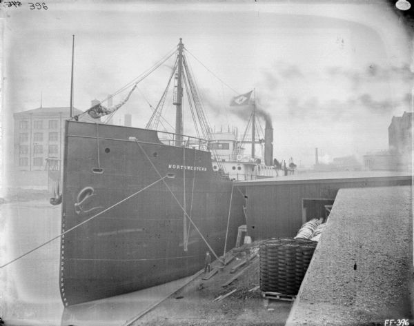 View looking down at ship at dock. A man stands at the edge of the dock just under the painted name of the ship, "Northwestern." Stacks of wheels are near a building on the right.