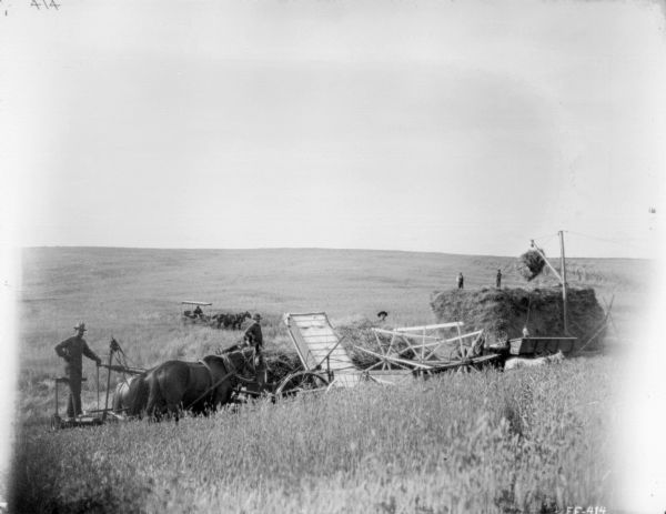 Elevated view of a group of men working in a field with a push binder header. On the far left a man is standing on a platform pulled by two horses. In the center two men are standing near the push header. On the right two men are standing on top of a large haystack, and below them at the base of the haystack another man is standing near a crane used to move headed grain to the top of the haystack. In the background is a man sitting in a horse-drawn buggy.