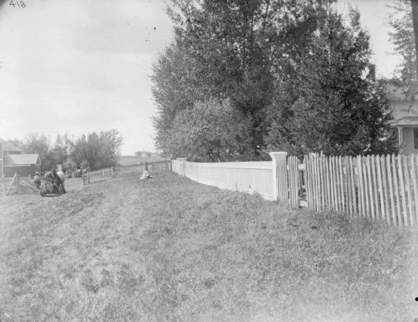 View across lawn towards a man, on the far left, riding a horse-drawn mower towards an open gate. A picket fence runs along the right side, with a farmhouse beyond. In the background in the center is a young girl sitting on the lawn in front of a fence enclosing the barnyard. There is a barn on the far left beyond the fence, and a man sitting in a horse-drawn buggy. In the far distance across a field is what appears to be a church.