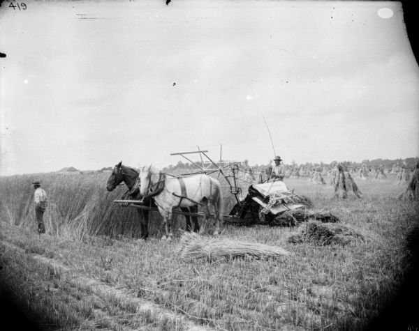 View across harvested section of field towards a man sitting on a horse-drawn McCormick binder. Another man is standing near the horses on the left. In the background across the field of tall grain are farm buildings. Shocks of harvested grain stand in the field in the background on the right.