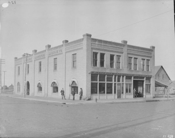 View across street towards the McCormick Harvesting Machine Company building at a street corner. Three men are standing on the sidewalk near the corner. Along the front of the building are show windows. Another man is standing near an open garage door on the right.