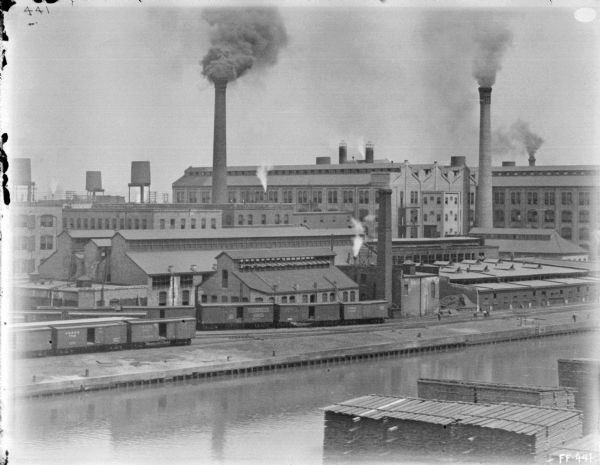 Elevated view across water towards McCormick Works. Lumber is stacked in the foreground. Along the far shoreline are railroad cars on railroad tracks. Smoke is billowing from a smokestack.