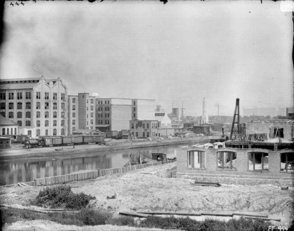 Elevated view from shoreline across canal towards McCormick Works. A building is partially constructed on the far right. There is a railroad train on railroads tracks along the far shoreline.