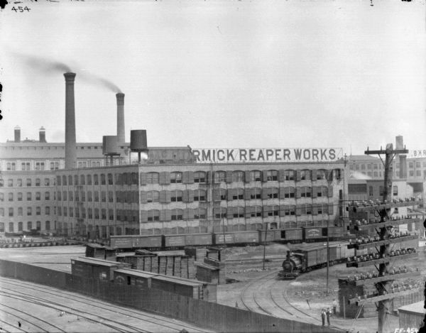 Elevated view of McCormick Works. In the foreground are powerlines, and beyond are railroad tracks and a locomotive pulling railroad cars. Near the factory are two smokestacks, and there are two water towers on the roof of one of the factory buildings.