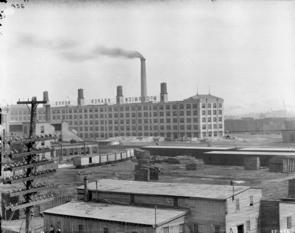 Elevated view of McCormick Works. In the foreground are powerlines, and beyond are railroad cars on railroad tracks. A large sign that reads (in reverse), "McCormick Reaper Works" is along the top of the factory building in the background near a smokestack.