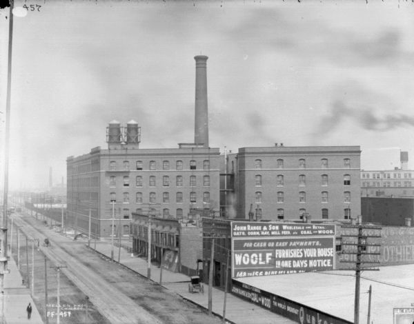 Elevated view across street of McCormick Works. In the foreground are sidewalks on either side of the street. Advertisements are painted on the sides of commercial buildings on the right.