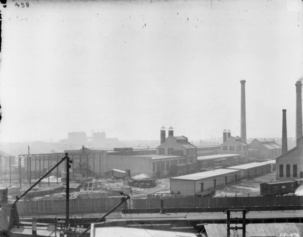 Elevated view of McCormick Works, with construction of wheel shop. In the foreground are the roofs of buildings, and what may be a ship on the lower left. There is a long wooden fence with railroad tracks and railroad cars. Factory buildings and a smokestack are beyond. Large buildings and what may be a bridge are in the far distance.