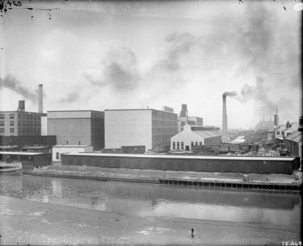 Elevated view across canal of the plant, with railroad boxcars behind a long, low industrial buildings along the far shoreline. Behind the railroad tracks are the factory buildings, with a smokestack on the far left. There appears to be a large church building in the far background on the right.