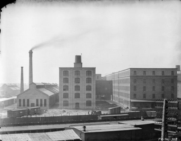 Elevated view of McCormick Works. Below in the foreground are low industrial buildings. A long platform with a fence is near railroad cars and railroad tracks. The plant yard is littered with parts and other equipment. In the background are factory buildings and smokestacks.