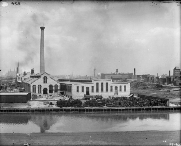 Elevated view across canal towards McCormick works. There are piles of parts along the shipping dock. A large church building is in the far distance.
