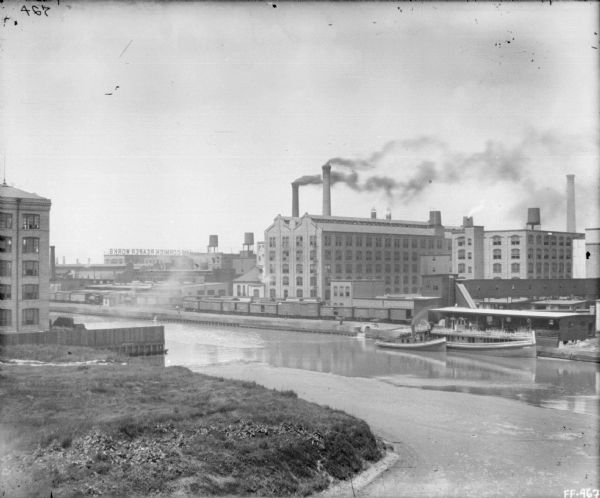 Elevated view over canal of docks and ships at the dock. In the background are railroad cars on a railroad track, factory buildings, and smokestacks.