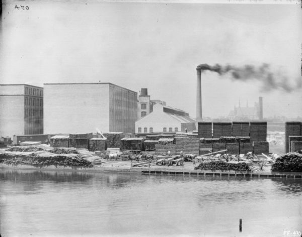 Elevated view across canal. Along the shoreline are piles of lumber along the shipping dock. Men are working with horse-drawn vehicles among the stacks of lumber. There is a large church building in the far background.
