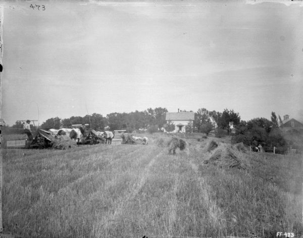 View across field towards three men working in a field with three binders. On the right a man is standing near fence with a group of cows. A group of people are sitting at the entrance to a farmhouse in the background among trees.