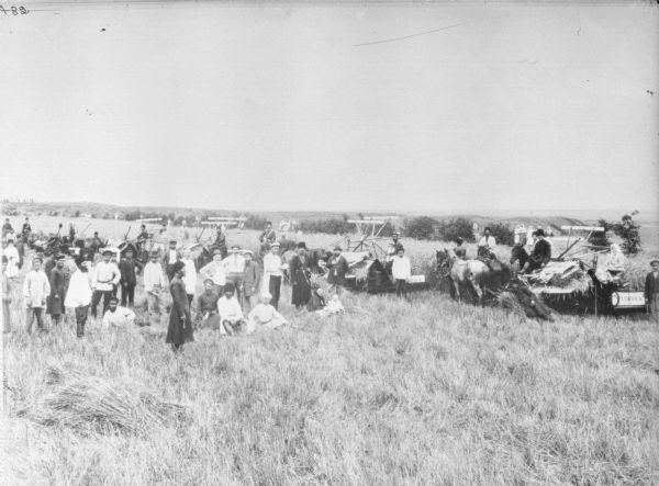 Slightly elevated view of a large group of people posing in a field. Men, women and children are posing near five horse-drawn McCormick binders. A group of women are standing together in the background on the right. In the far background is a valley.