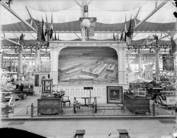 View of display of machines and equipment at the booth for the McCormick Harvesting Machine Co." In the center is a framed display of "The Great McCormick Reaper Works in Miniature." A framed portrait of Cyrus McCormick is on the floor in front. Small models of a reaper and mower are on display tables on the left and right flanking the booth. Flags are at the top of the framed display, and an iron gate is along the front.