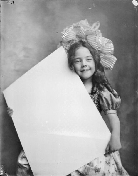 A young girl wearing a bonnet and a dress is sitting and holding a large, blank, white piece of paper in a studio.