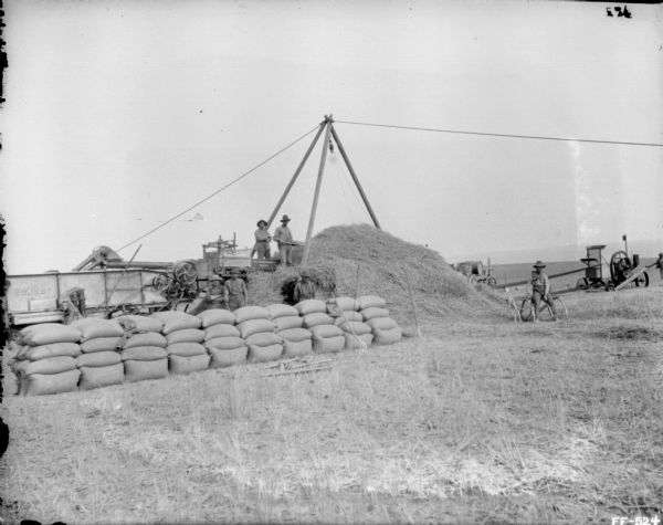 View across field towards piles of grain in bags stacked near a harvesting operation. Men are standing near a large haystack. On the far right is a tractor providing belt-driven power to the machinery.