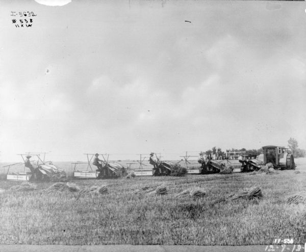 View across harvested field towards five men on five McCormick binders being drawn by a tractor.