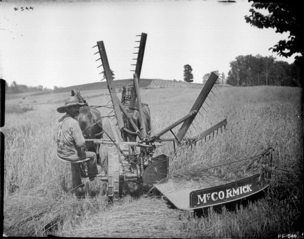 Rear view of a man, sitting and turning to look over his right shoulder, on a horse-drawn McCormick reaper in a field.