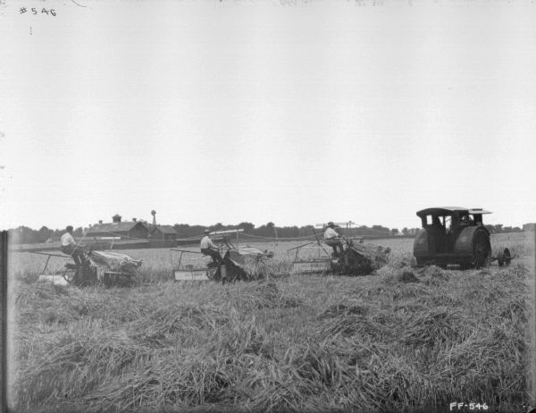 View across field towards three men on three McCormick reapers drawn by a tractor. Farm buildings are in the background on the left.