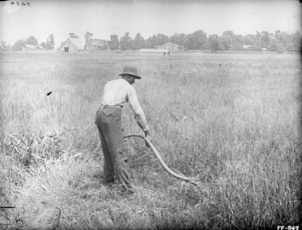 A man is working in a field using a scythe. In the background are farm buildings.